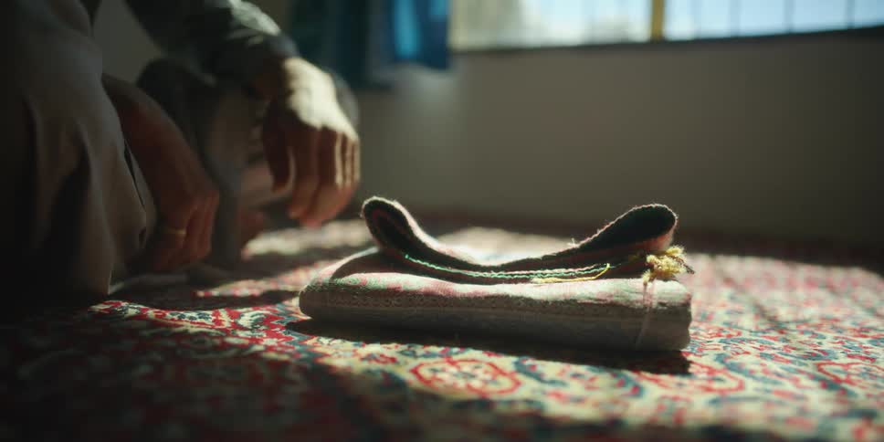Person drops matt onto the floor in square of sunlight steaming through the window with blue curtains then sits with legs crossed beside it outside of the light and plays with the ring on their pinky finger