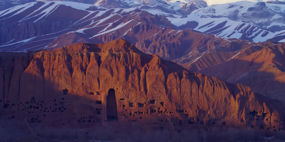 Shadows fall across parts of cliff dwelling on mountain in mountain range that is partially snow covered