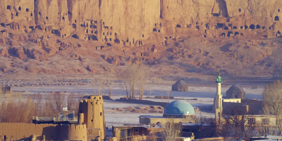 Buildings made of various materials and sizes sit near a canyon where primitive homes are carved into the side