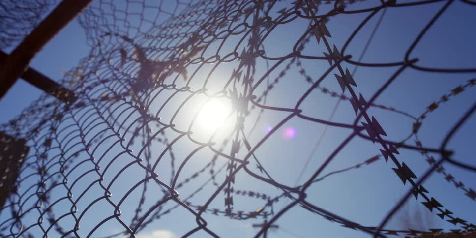 A fence with barbed wire with the sun the sky and clouds above