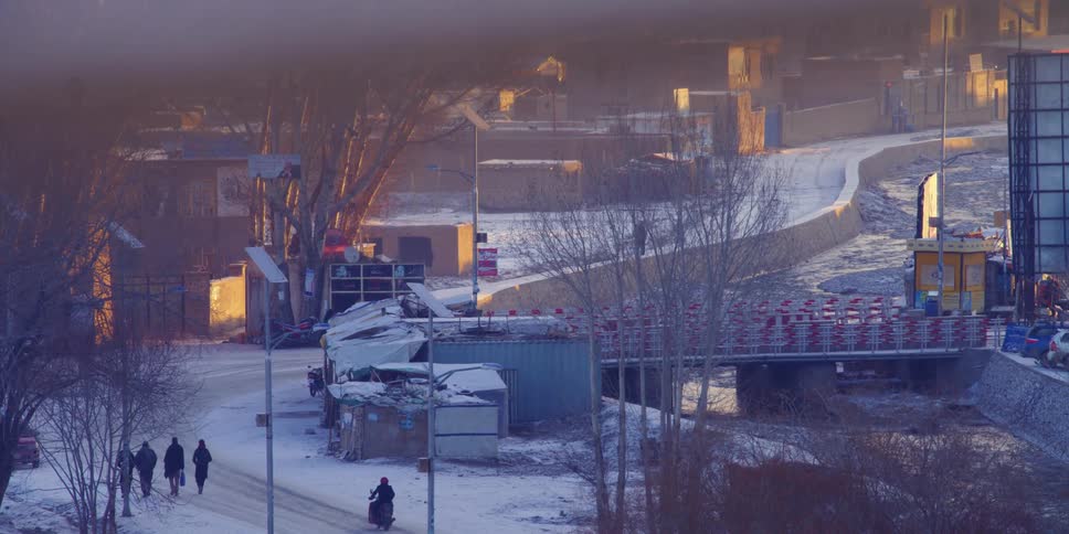 Pedestrians and small vehicles travel along a snowy road running alongside a river through a town during the day