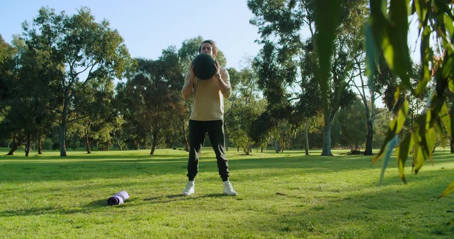 A man does squats with an exercise ball in a field on a sunny day