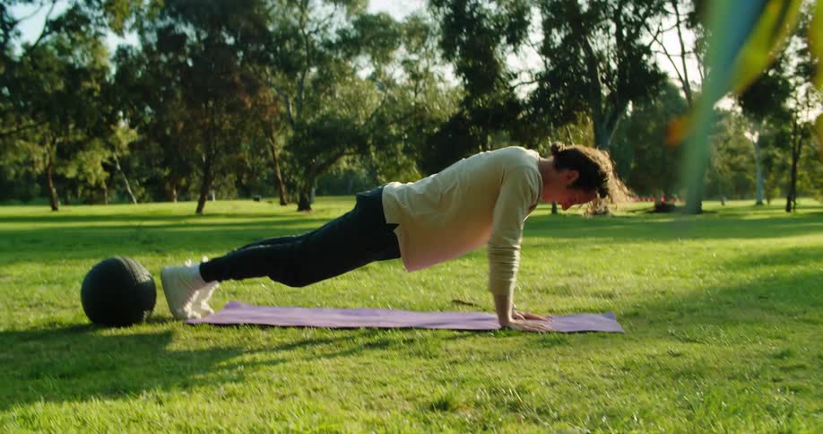 Man with long hair a brown sweatshirt and black track pants does push ups on the lawn in the park