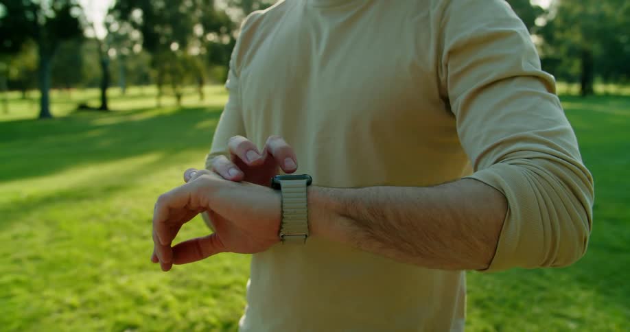 Man with long hair and a brown sweatshirt uses a smartwatch on the lawn in public park