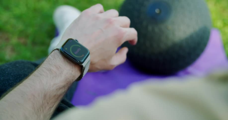 Person sitting on purple mat on grass near ball adjusts smartwatch