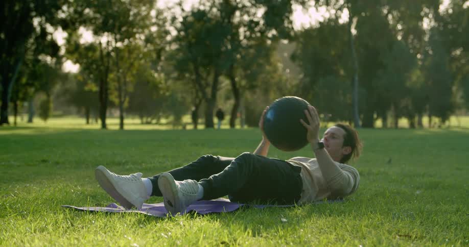 A man does sit ups in a public park while holding an exercise ball