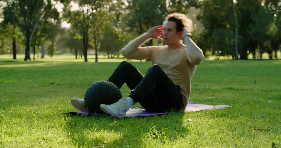 A man does sit ups on a mat in a field in a park on a sunny day