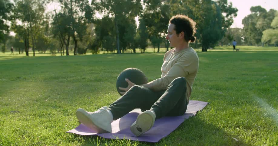A man sitting on a mat on grass and using an exercise ball