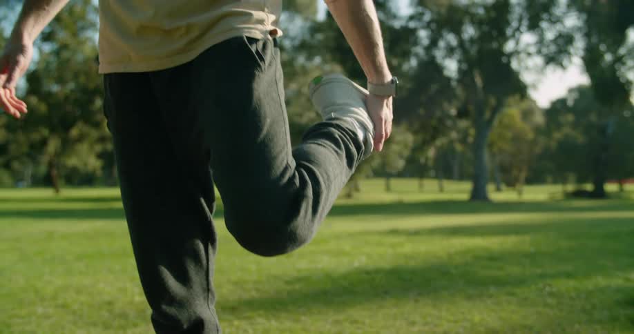 Man with a brown sweatshirt and black track pants warms up before working out on the lawn in the public park