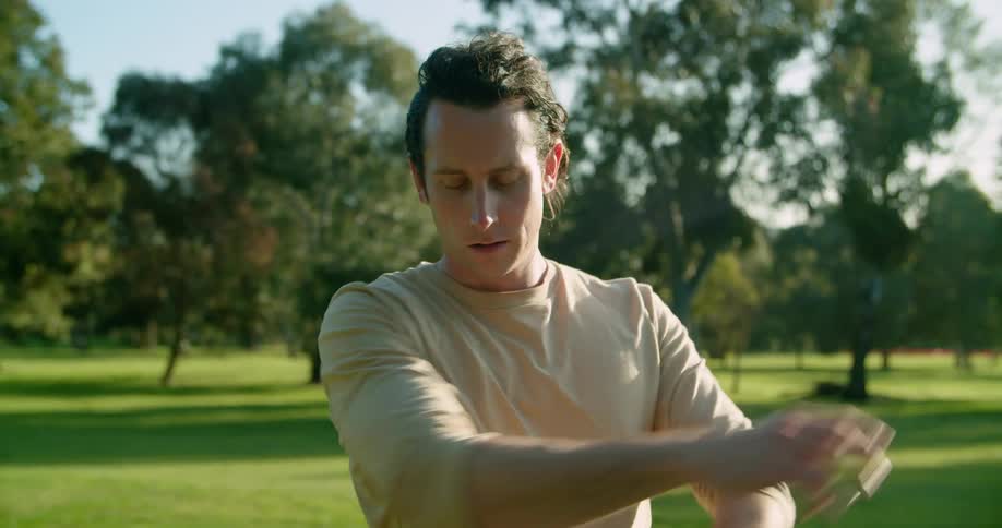 Man with long hair and a brown sweatshirt warms up before working out on green grass in the public park