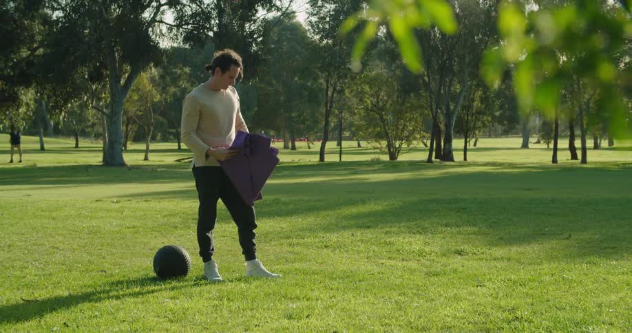Young man places an exercise mat and a weighted ball over the grass in a park