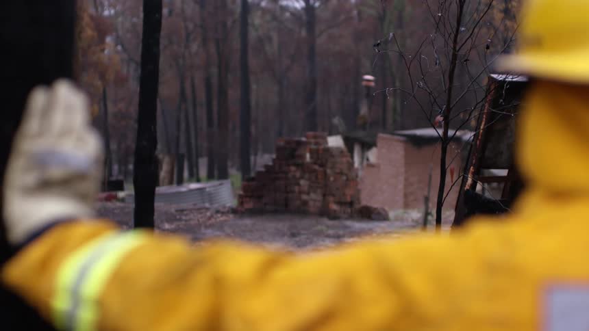 A firefighter looks at the ruins of a building in a woodland while resting a hand against a tree
