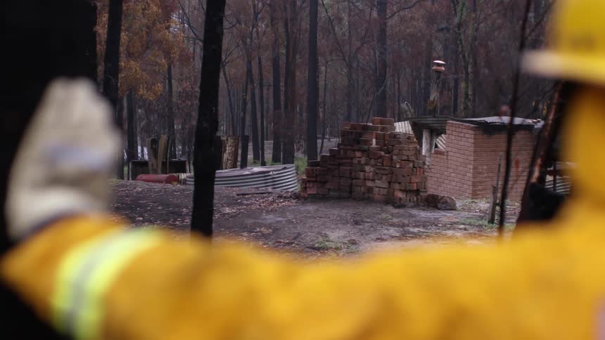 Person wearing firefighter protective gear leans hand against tree looking toward water and structure near bricks