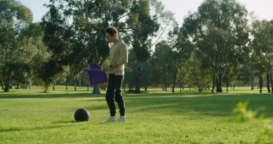 A man carries a yoga mat and exercise ball and places them on the ground in a public park open grass