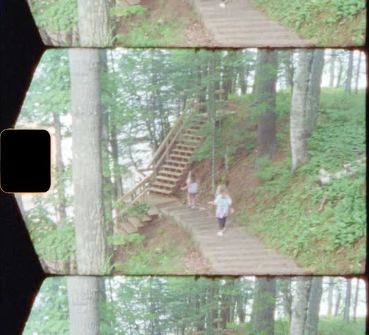 A pair of girls descend a cement staircase on the side of a hill in a woodland during the day toward lake 8mm