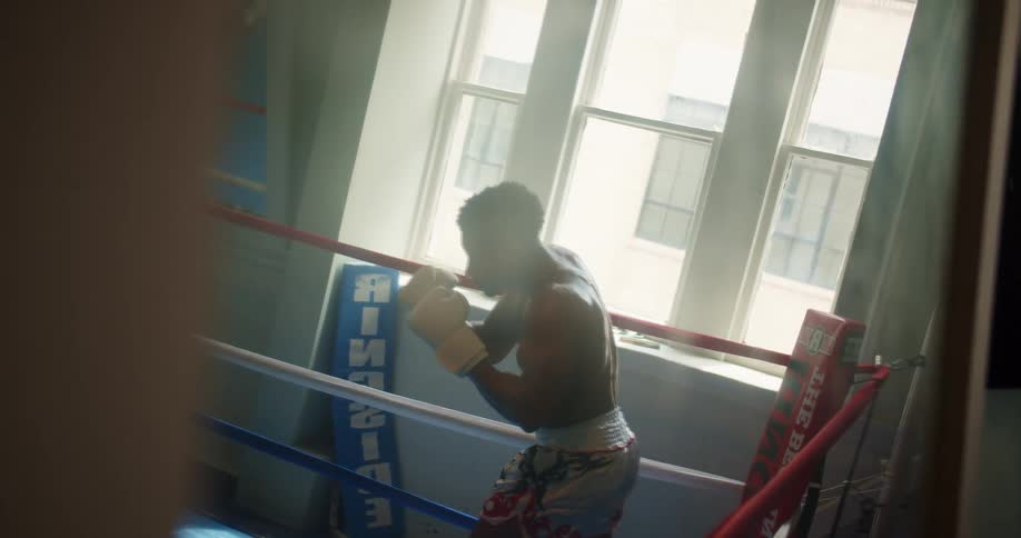 Man shadow boxes standing in boxing ring near window showing another building