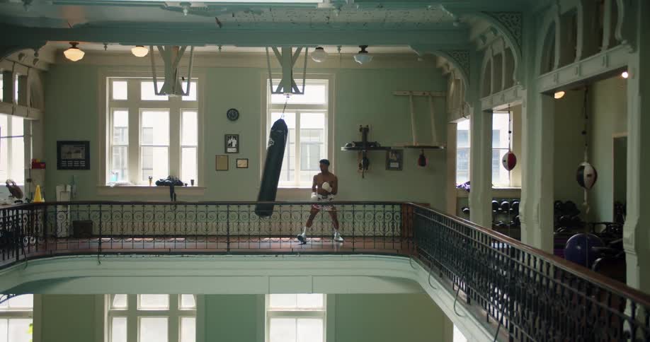 Man in boxing gloves and shorts punches heavy bag on balcony in gym