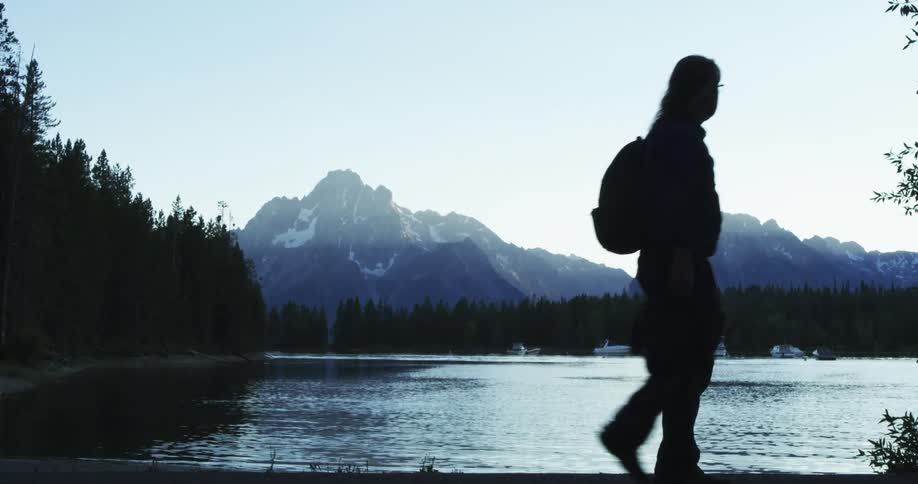 Man wearing backpack walks next to a lake near the pine forest on the mountains at sunset
