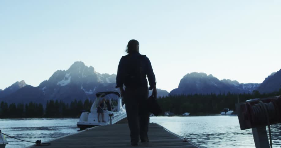 A man walks down a dock towards a woman standing on the back of a boat