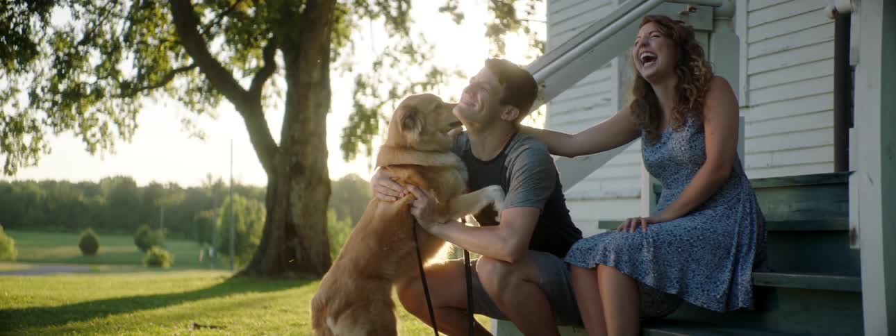 A man and woman laugh as they sit on their porch with their dog and he jumps up for a treat