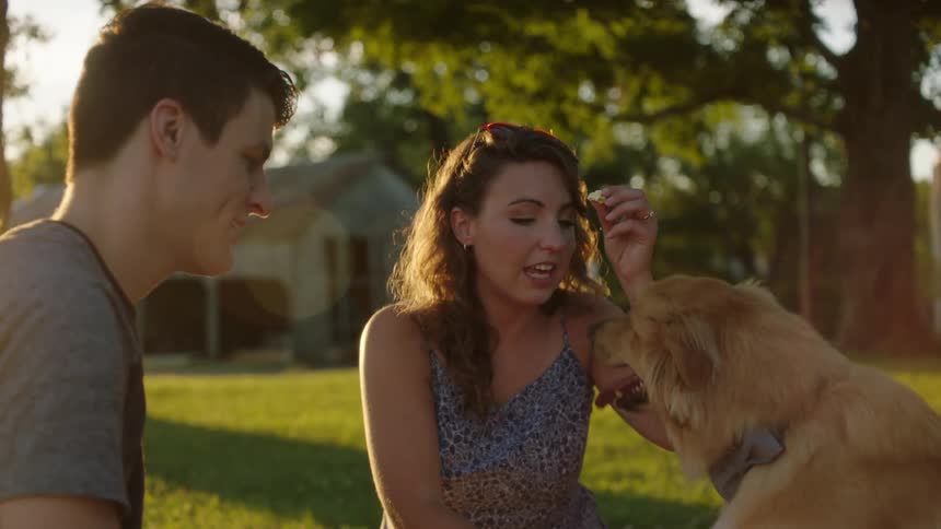 A man and a woman sitting on grass feeding popcorn to a dog