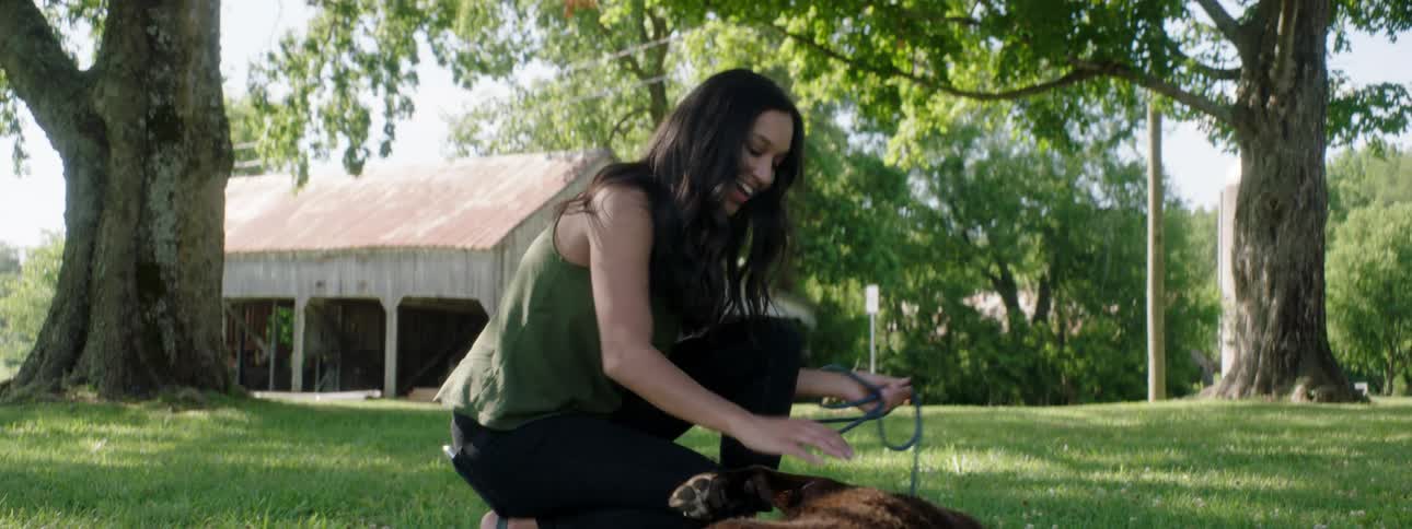 A woman bends down and plays with a dog in a rural grass area with worn barn in the background