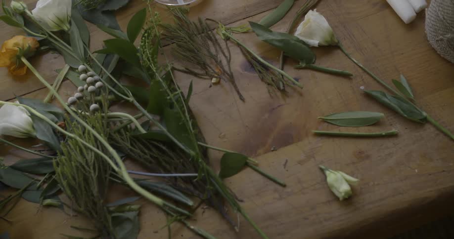 A woman picking up a pair of shears from a table littered with flowers and foliage
