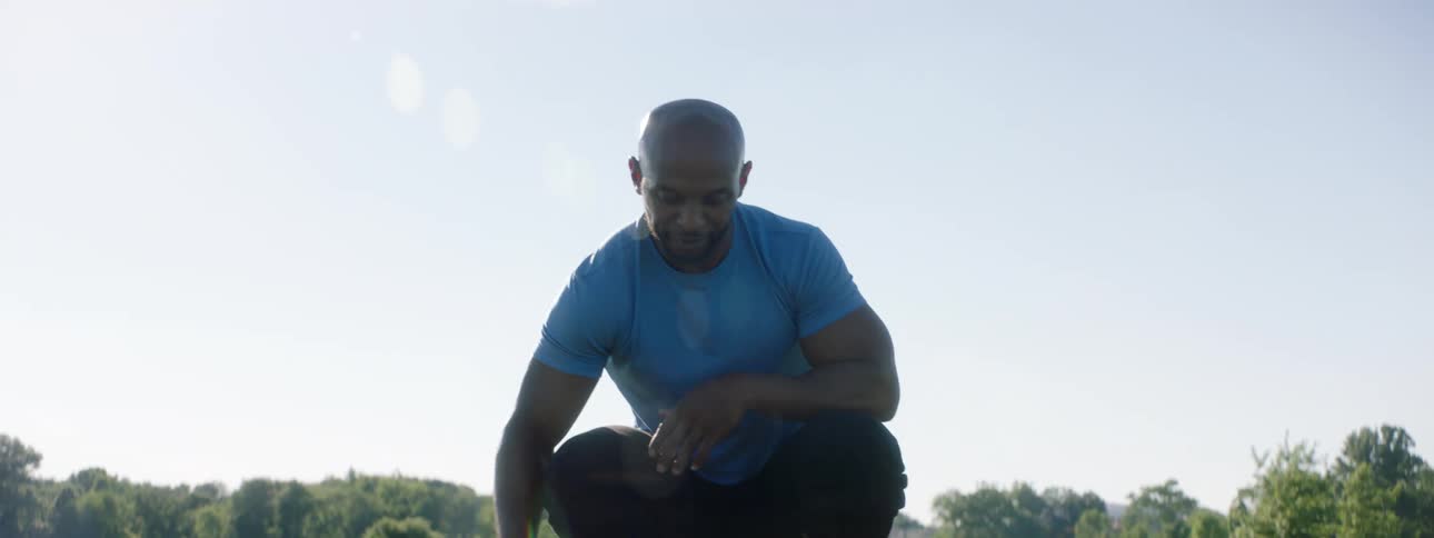Bald man with a blue t shirt and black pants pets a brown dog on green grass in the field on a sunny day