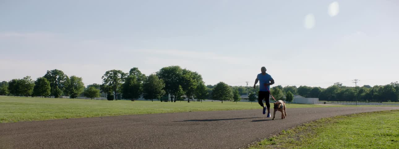 Athletic man with a pet dog on a leash jogging on a paved road in a park at day