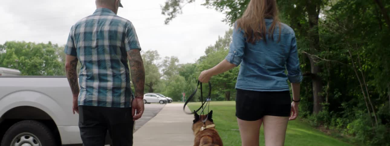 A man and woman walk their dog on a sidewalk beside commercial truck