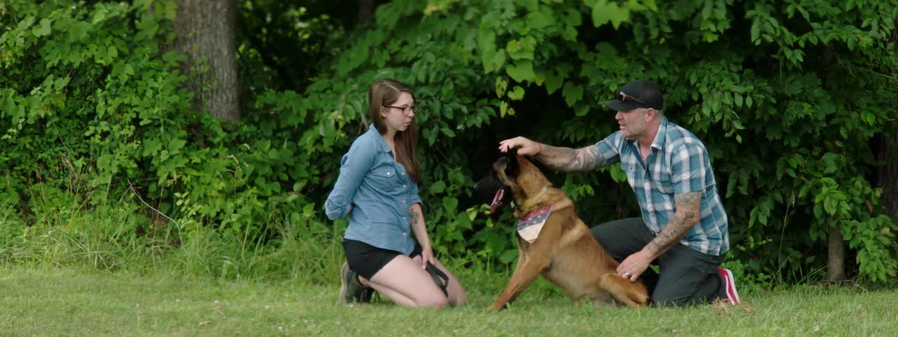 A man kneeling on the ground pets a dog and talks to a woman kneeling in front of the dog holding his leash