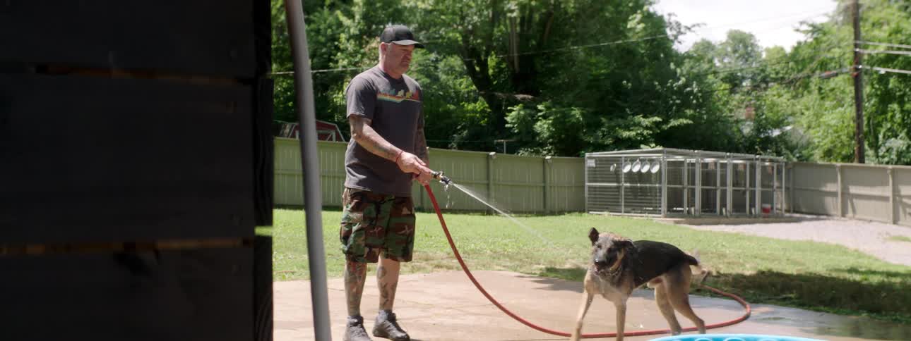 A man with a hat spraying a dog with a hose