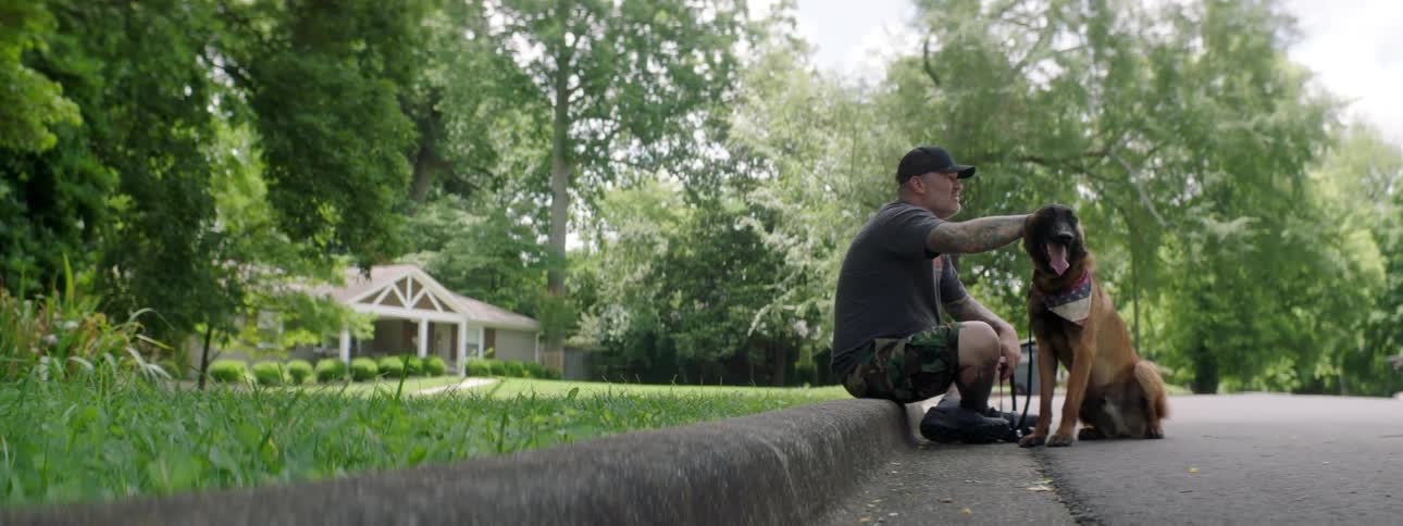 Tattooed man with a cap a t shirt and shorts sits on the sidewalk and pets a brown dog