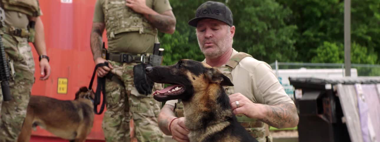 A kneeling soldier pets a dog while talking with other soldiers standing nearby
