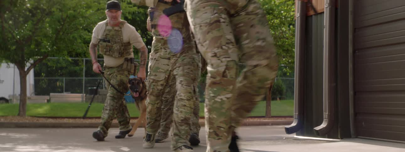 Four men in army uniforms go around the corner of a building with guns raised a dog being led