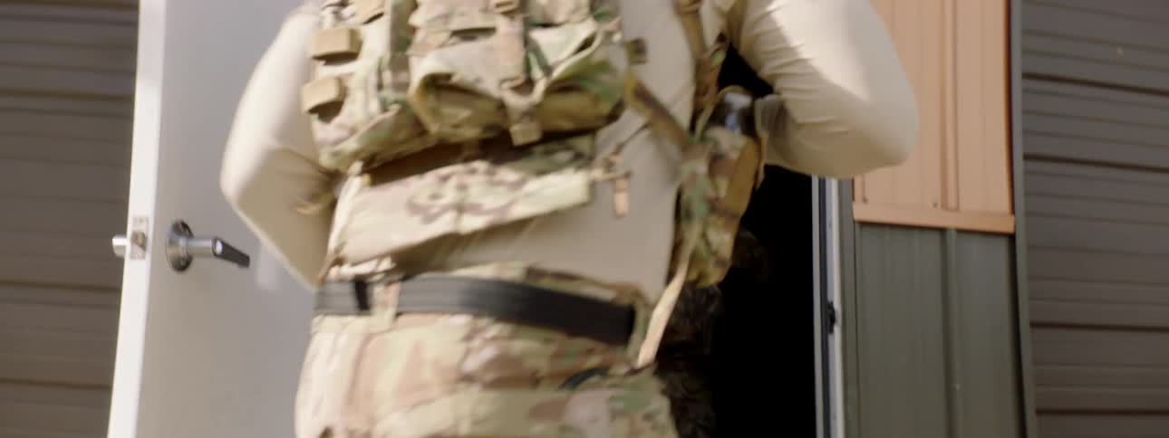 A military officer in camouflage holds open the door to a building for fellow military officers with guns drawn