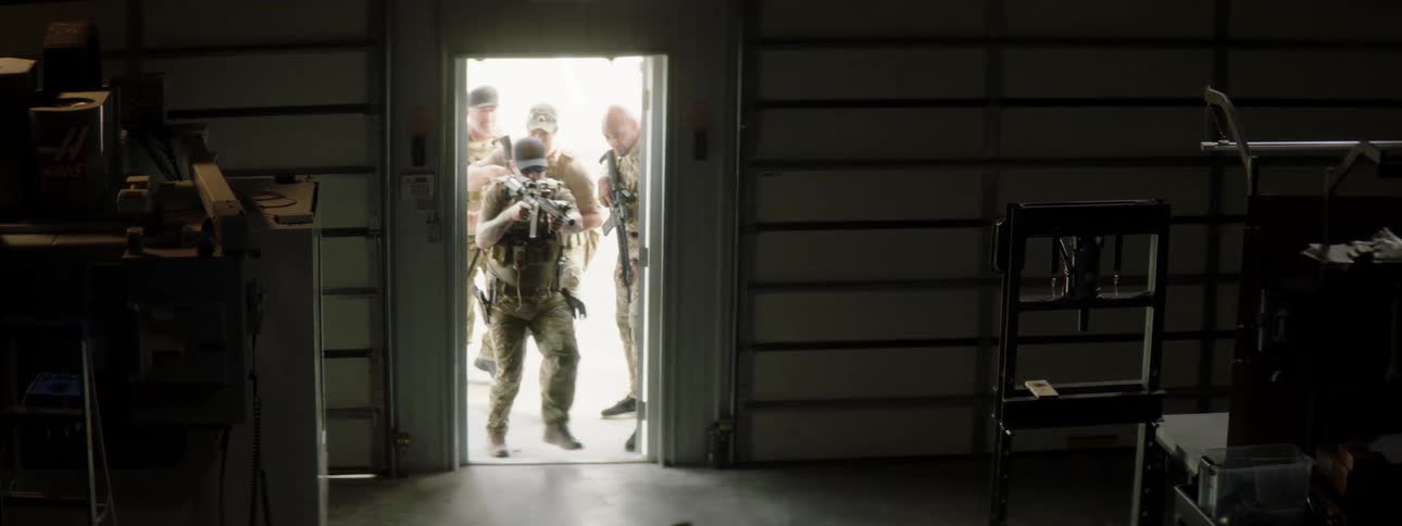 Multiple military men and their dog walk into a large shed with their weapons drawn
