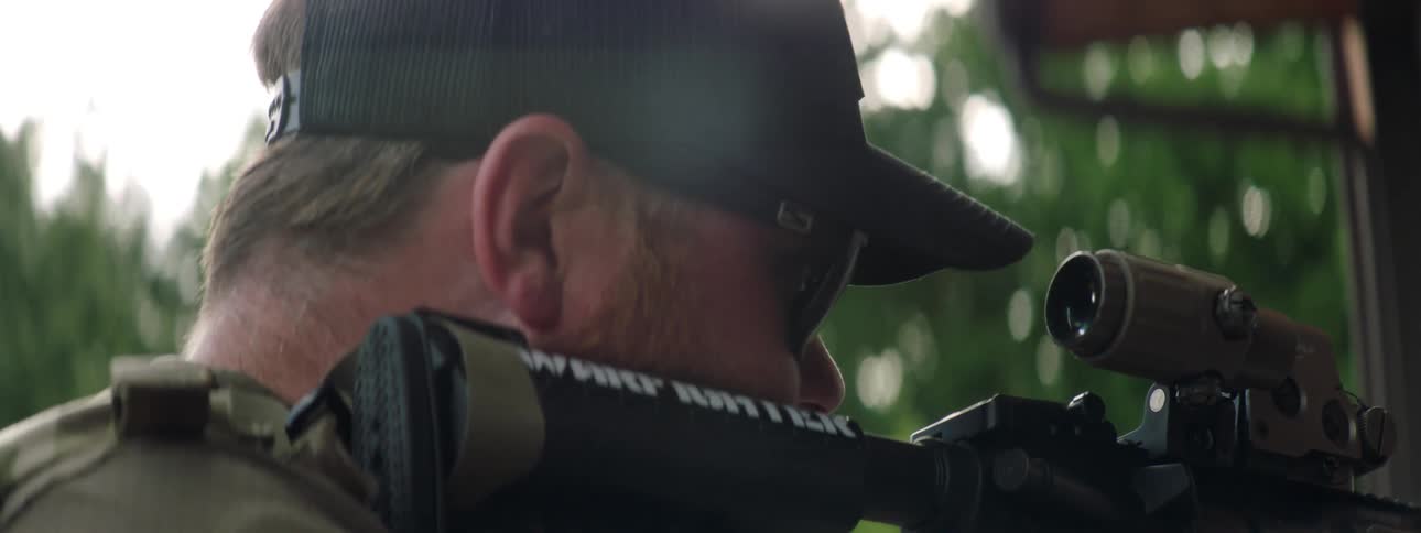 A military officer stands outside a home looking through the scope of a rifle on his shoulder