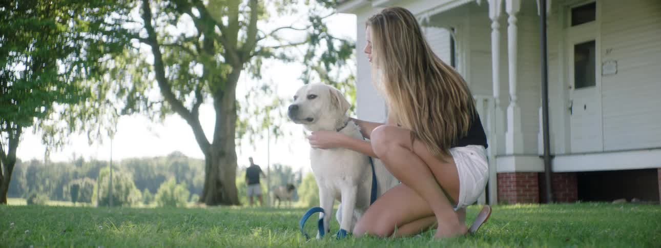 A young female crouches down and pets her dog in the shade of a tree