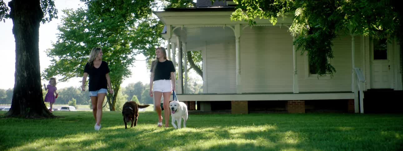 Two women walk their dogs in a neighborhood as a little girl plays outside a house