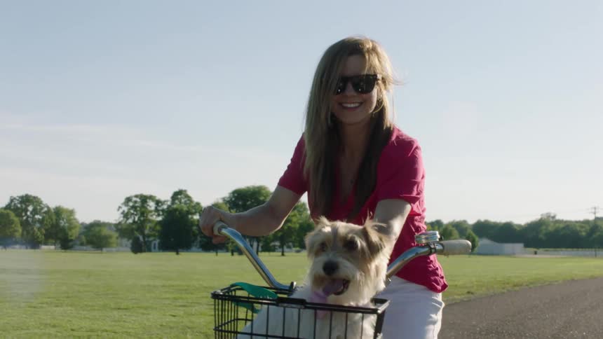 A young woman rides a bike on a rural gravel pathway with a dog in the basket