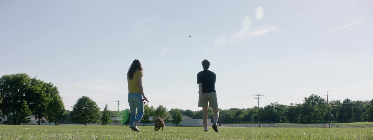 A man and woman stand in a large grassy park area and throw a ball for their dog to fetch