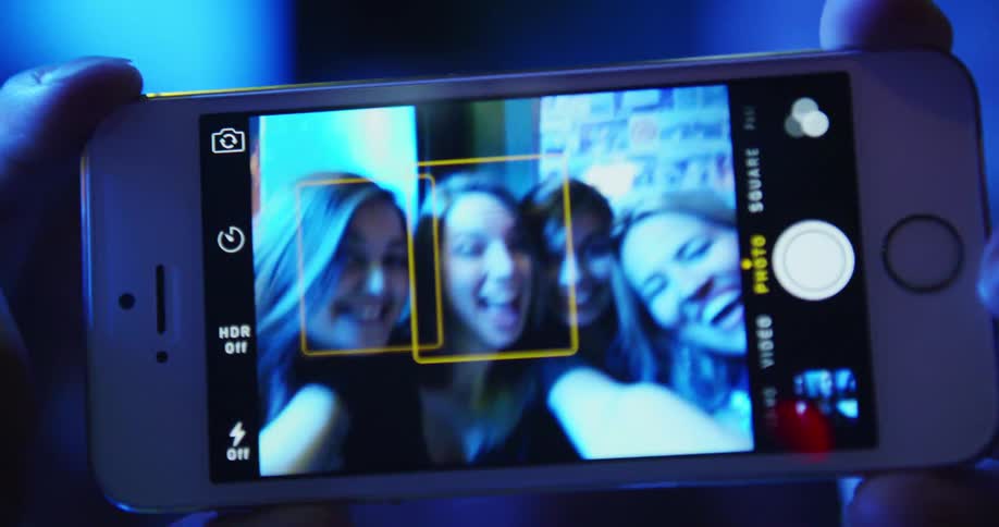 A group of women pose together as one holds a smartphone in preparation for a selfie