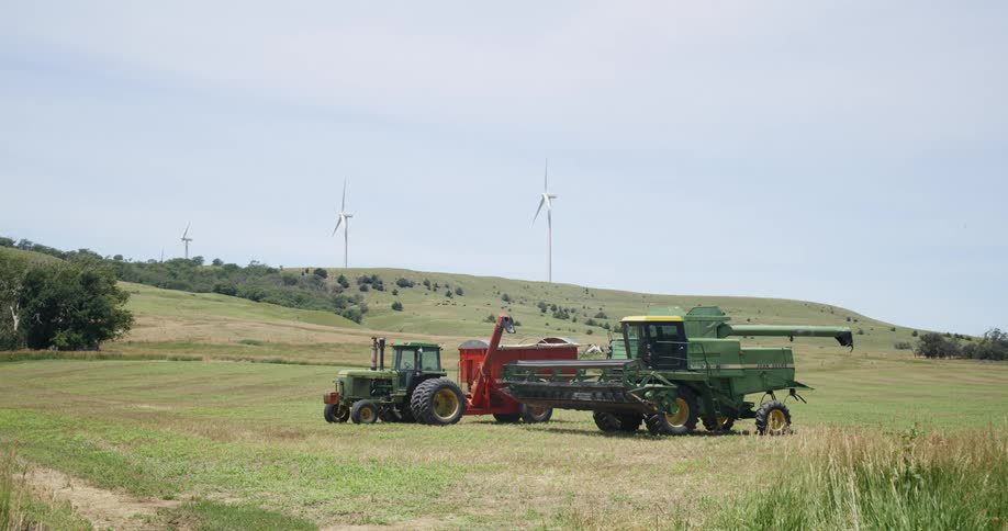 A tractor and a harvester sitting in a field near a wind farm on a sunny day