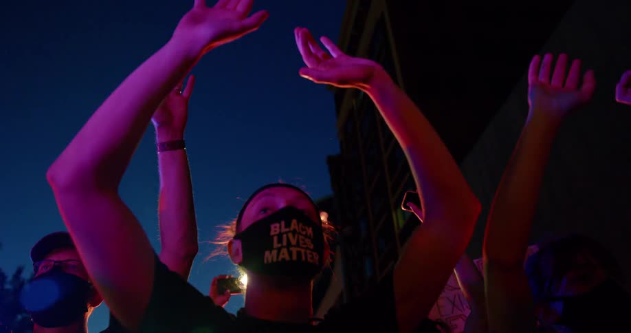 Black lives matter protesters stand outside at night with their arms raised as police lights flash around them