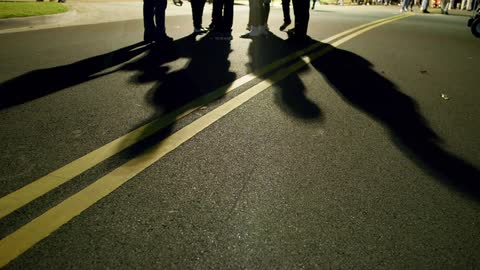 People stand and walk along asphalt road as their shadows cross double yellow line