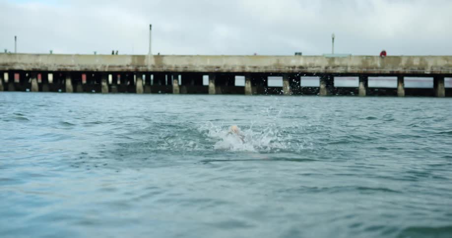 A man swims in a large calm body of water with a pier or bridge in the background