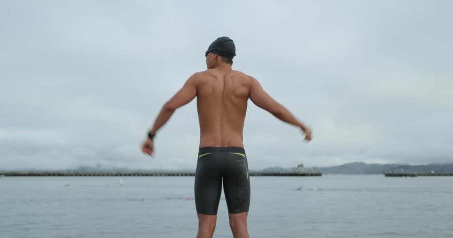 A man wearing a swim cap and spandex shorts stretches in front of the ocean as dolphins swim nearby