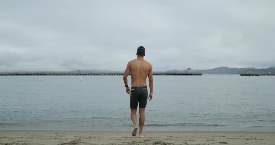 A man with a swimming cap and goggles walking on a beach into the water