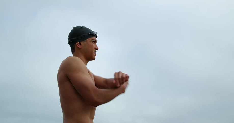 A man with goggles and a swimming cap stretching with the sky and clouds in the background